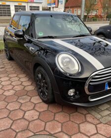 Ein schwarzer Mini Cooper mit weißen Streifen auf der Haube steht auf einem Parkplatz