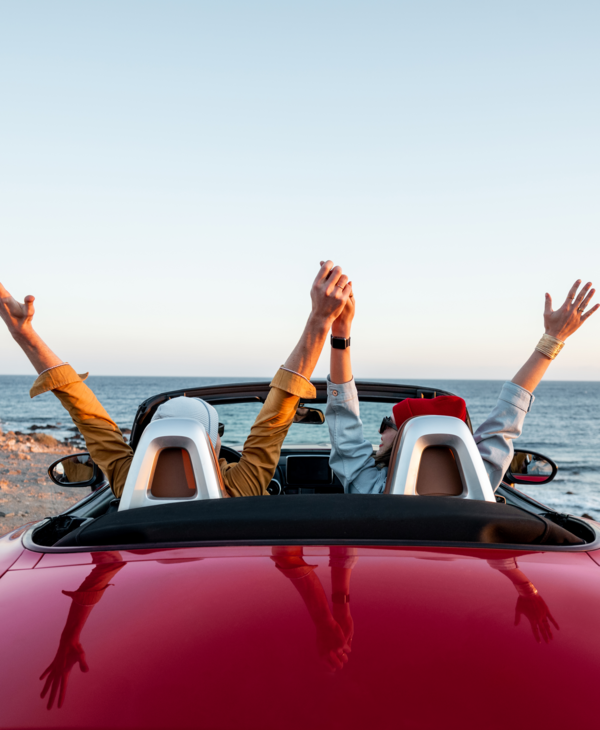 Zwei Personen sitzen jubelnd in einem roten Cabrio und parken am Strand